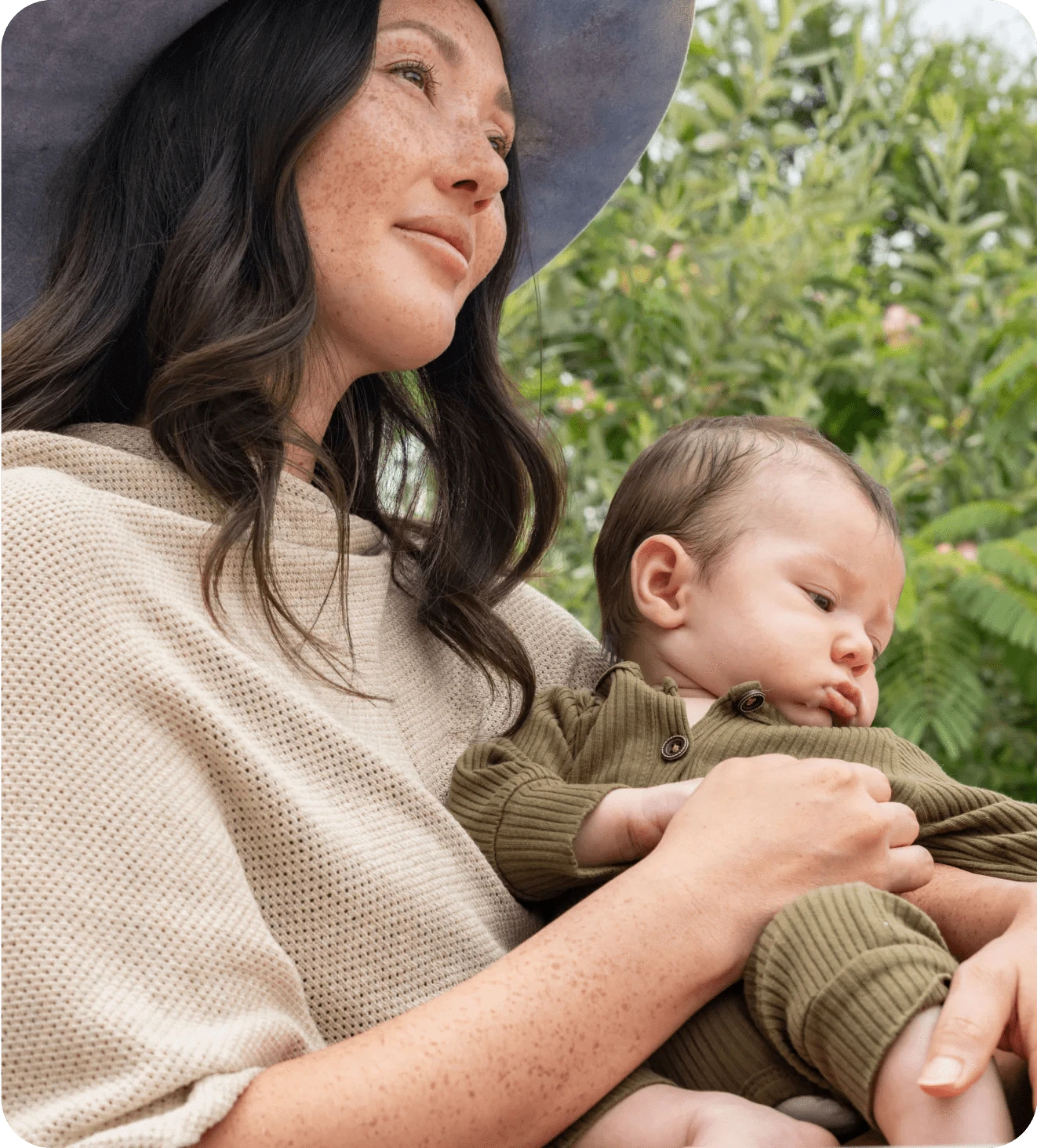 A woman in a hat lovingly holds an infant outdoors.
