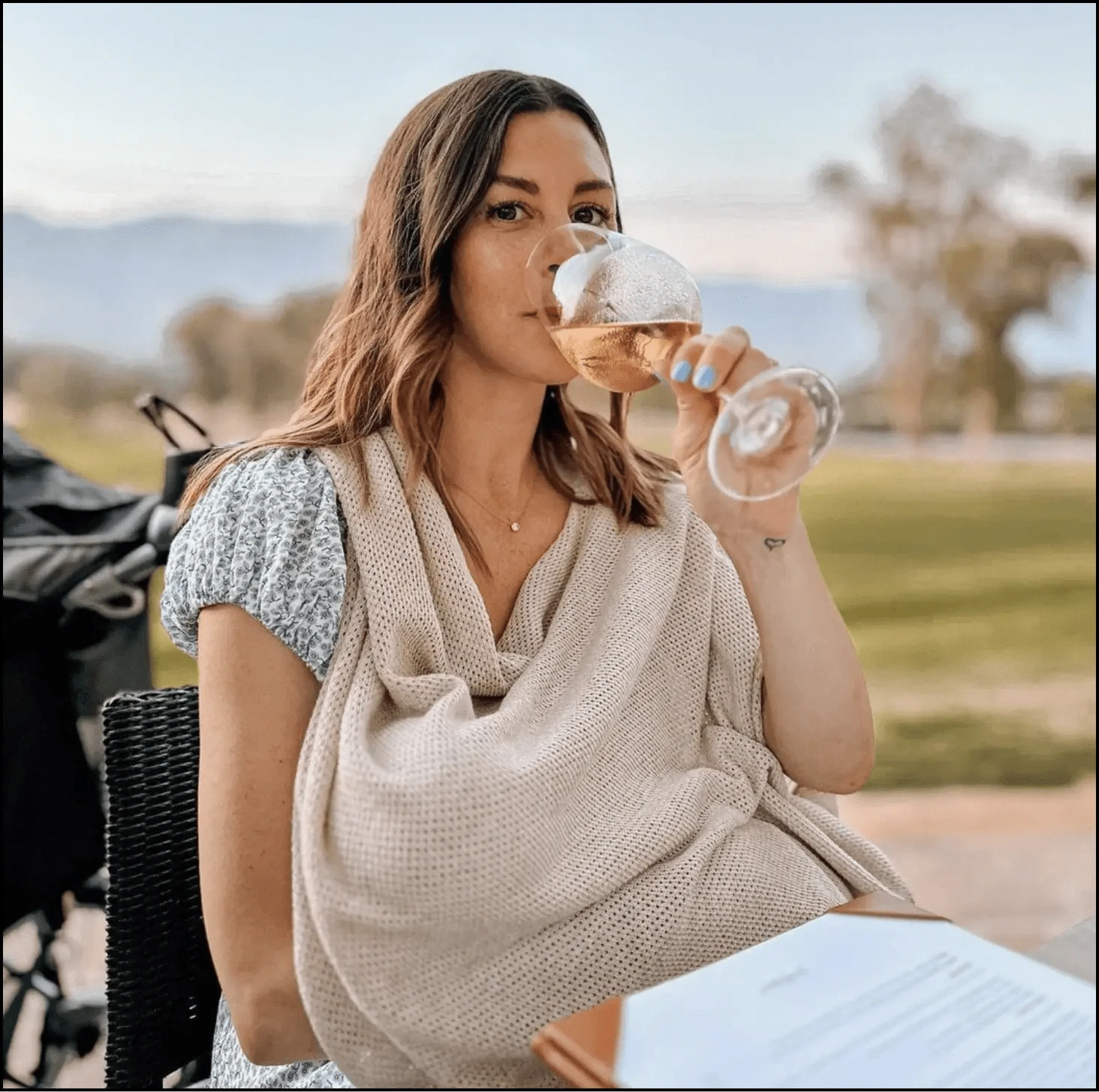 Woman sitting outdoors, sipping wine with a serene background.