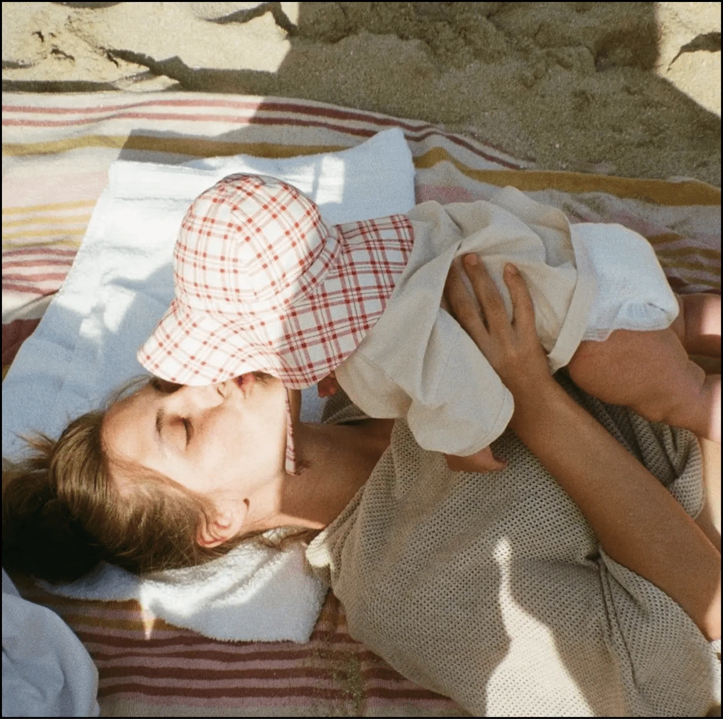 Woman lying on a beach towel, kissing a baby wearing a plaid sun hat.