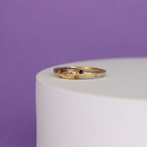 product shot of 9k yellow gold ring with sapphire gemstones sitting on white plinth against a purple background 