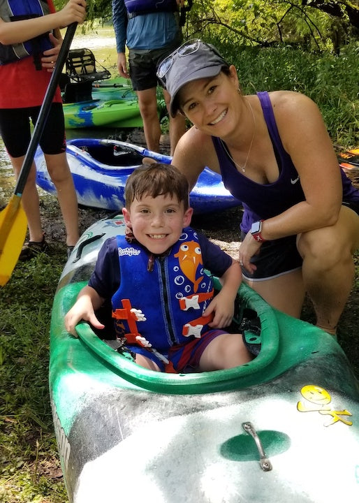 smiling little boy sitting in a kayak onshore; woman kneeling next to him