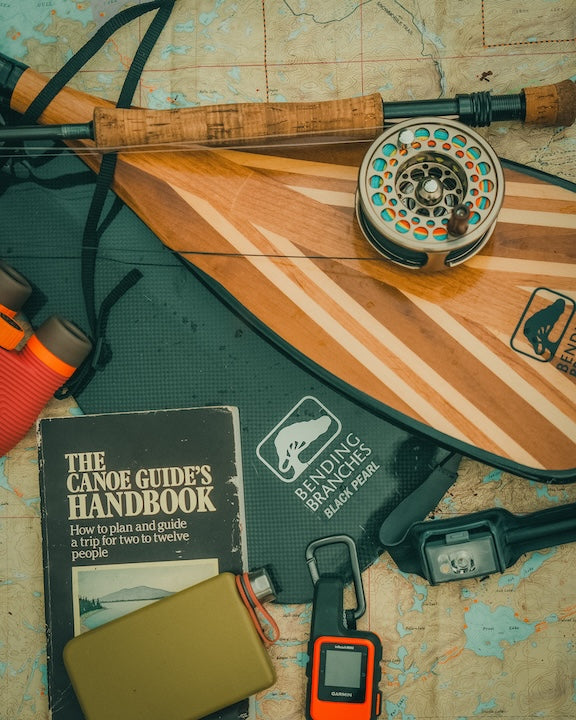 Branches paddle, fly rod, canoe handbook and other gadgets laying on a Boundary Waters map