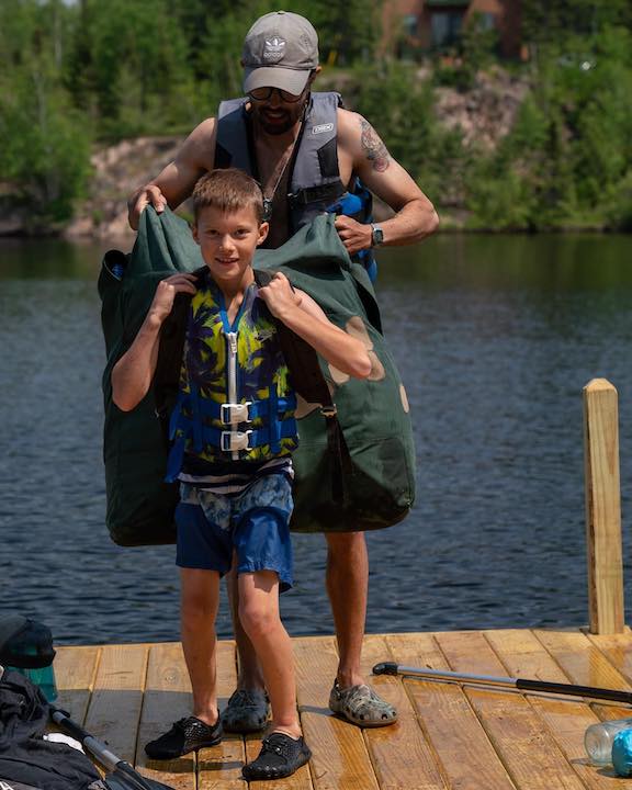 counselor helps a young boy put on a canoe pack on the dock