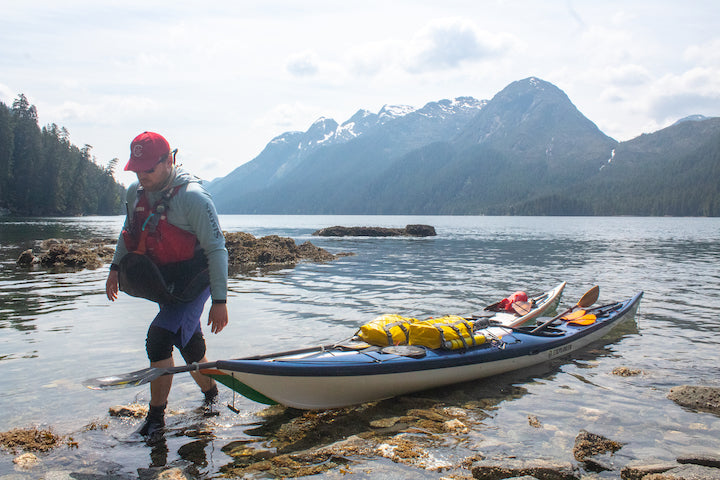 Nuka walks on-shore next to kayaks, Alaskan mountains in the background