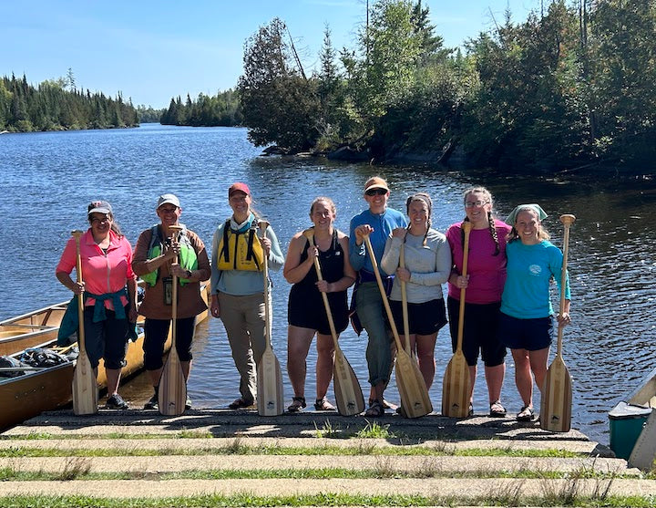 8 women stand at lake's edge with paddles and canoes