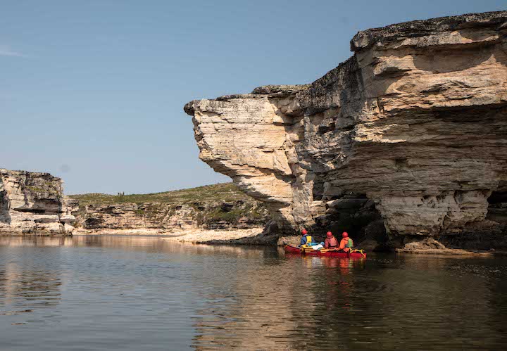 canoe with three people paddle by a shoreline cliff