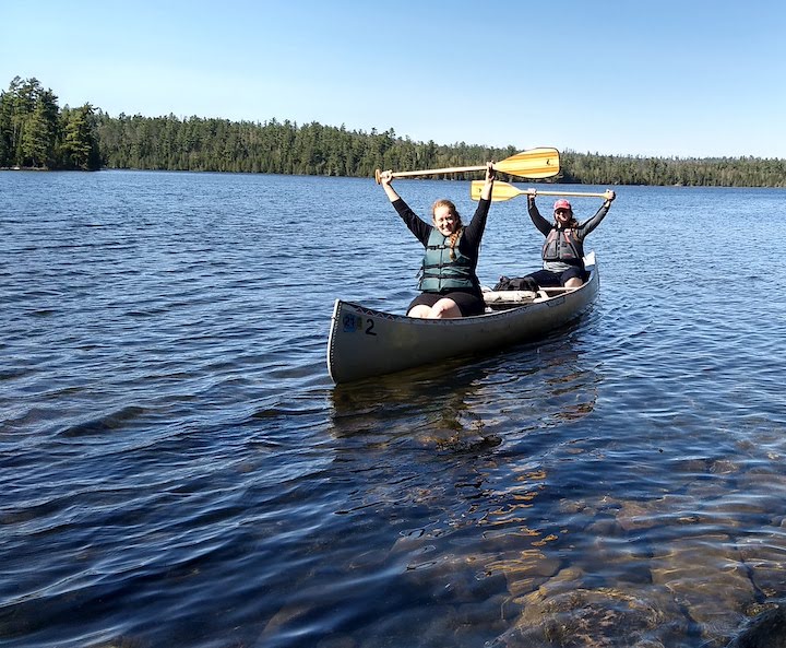 two women in a canoe on a lake, raising their canoe paddles high