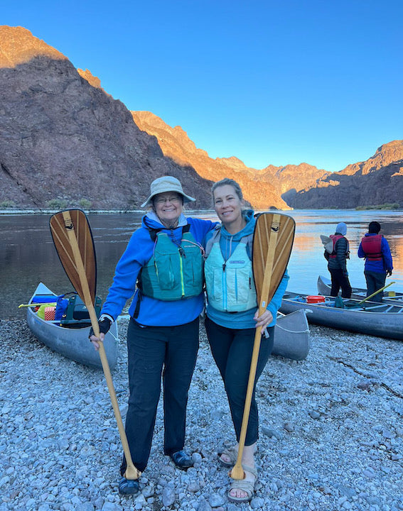 Kristen Elgo (right) with another Girl Scouts volunteer, standing with their Bending Branches paddles in front of canoes and next to the Colorado River