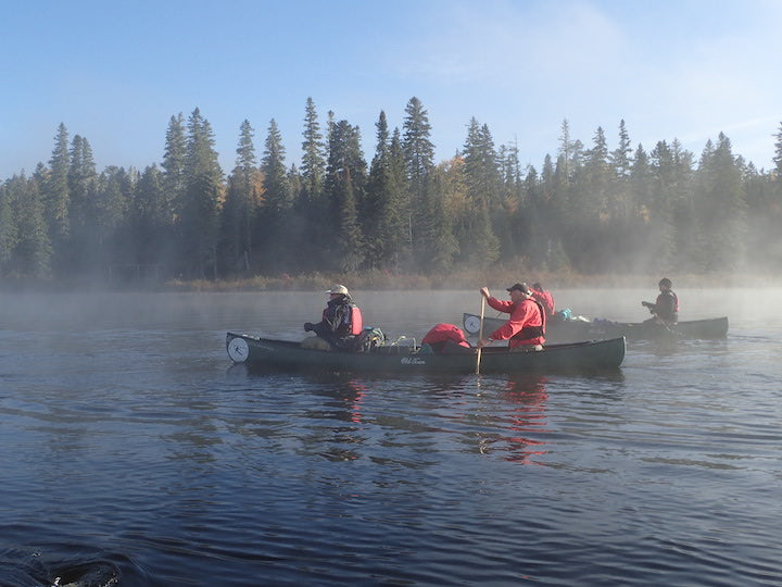 men in two canoes paddling on a lake in the mist, pine trees on the shore