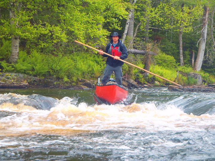 man  standing in a canoe, poling down some river rapids