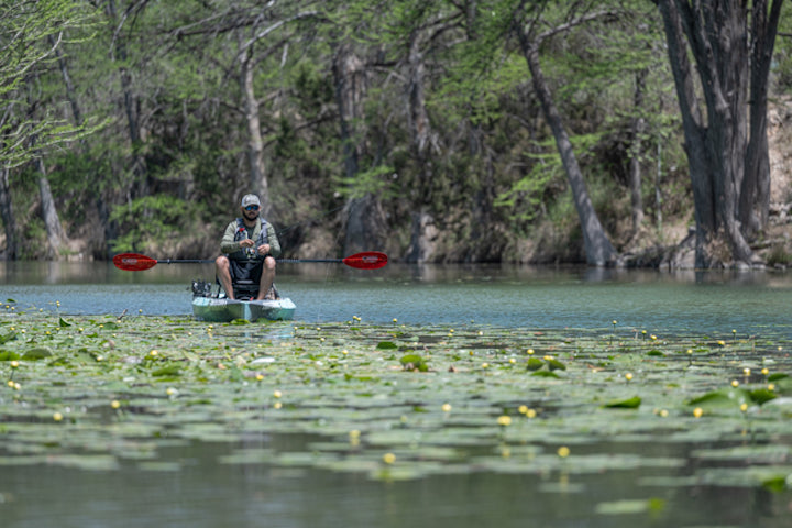 a man kayak fishes in a river among lily pads