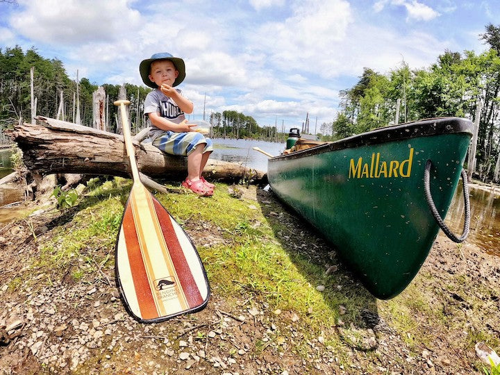 A little boy sits on a log next to his canoe and a Bending Branches paddle at the lakeshore