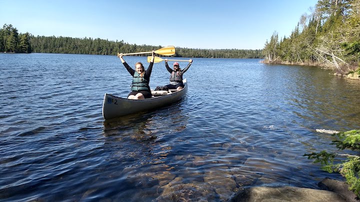 two women in a canoe with paddles raised, near a portage