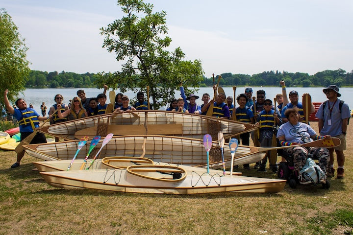A large group of people next to a lake with several canoes and kayaks built through Urban Boatbuilders