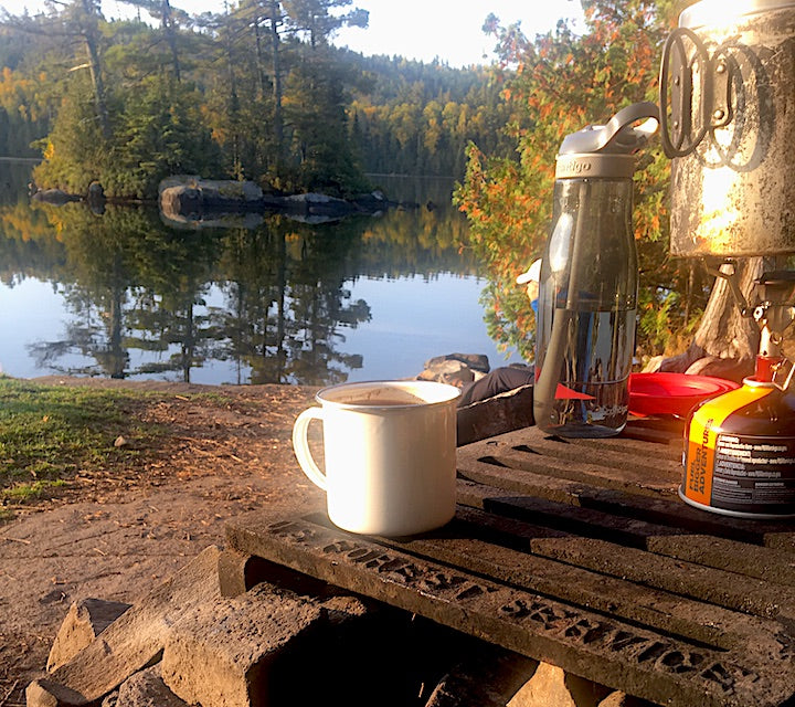 cup of coffee and water bottle next to camp stove at a canoe campsite