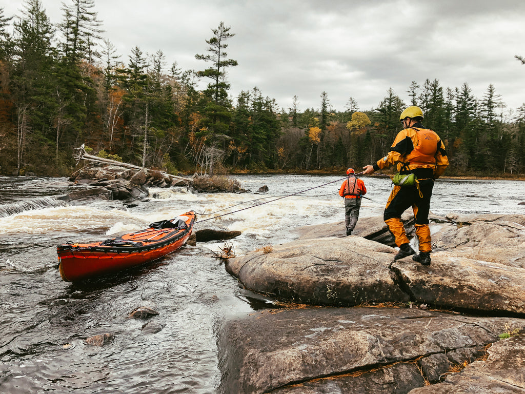 Two men are wearing a dry suits and holding rope tied to a red canoe in the water, lining the boat down a rapid on the Dumoine River.