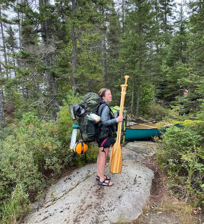 Woman standing along shore with two packs, carrying canoe paddles