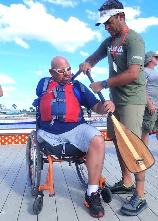 a man helps a wheelchair-bound man with the one-arm freedom canoe paddle