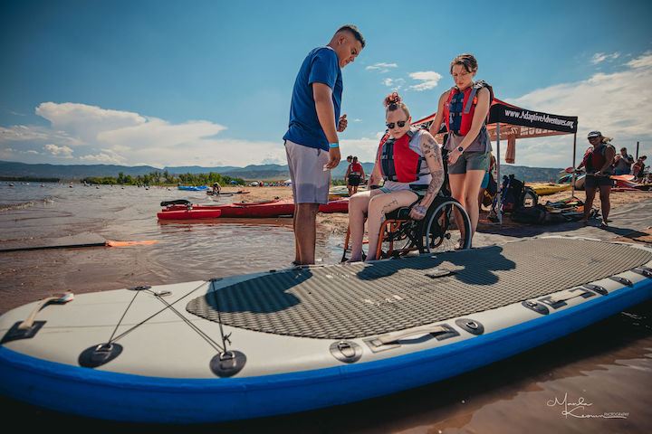 man and woman help another woman in a wheelchair get on a paddleboard