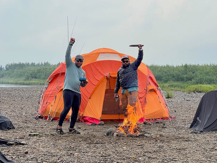 two men stand in front of an orange tent on the beach