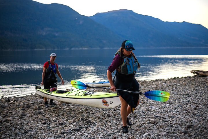 woman and man carrying a kayak up on the beach from the water, mountains in the background