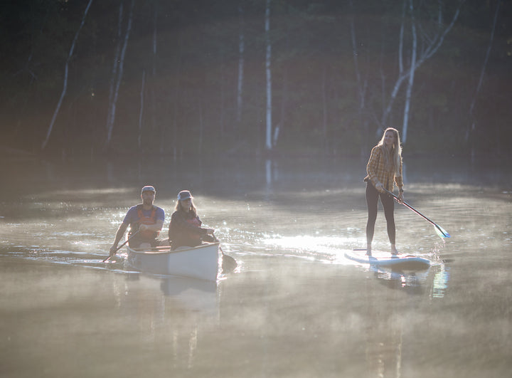 two people in a canoe next two a woman on a paddle board on a calm misty lake