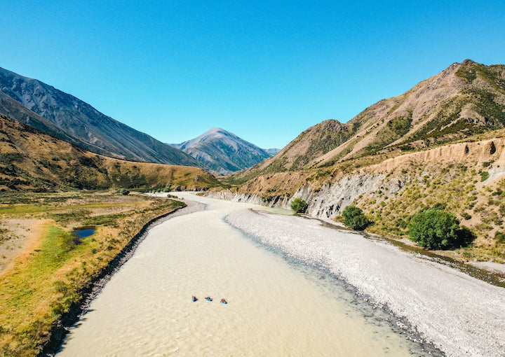 three packrafters in the distance on a silty river in the mountains of New Zealand