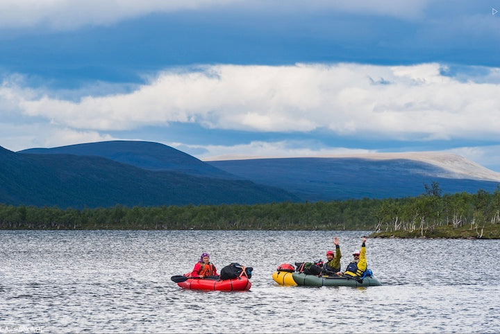 three packrafters on the water waving, with mountains in the background