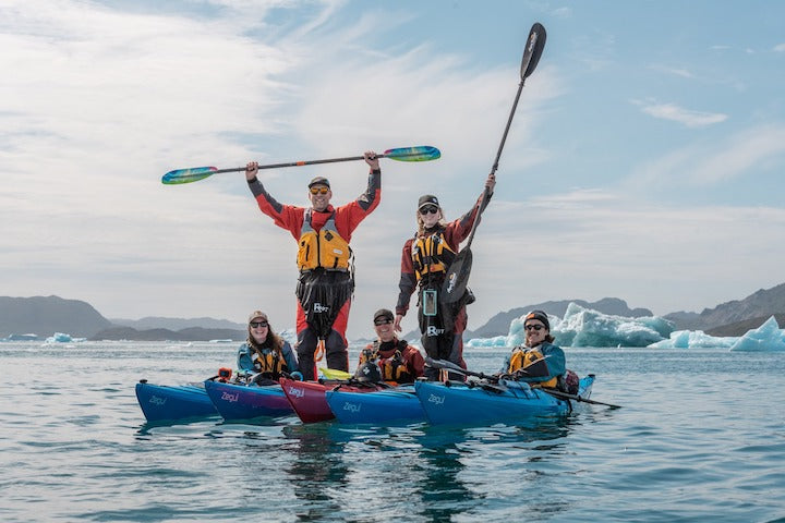 three people in sea kayaks while two stand up in their kayaks, on the water with icebergs in the background