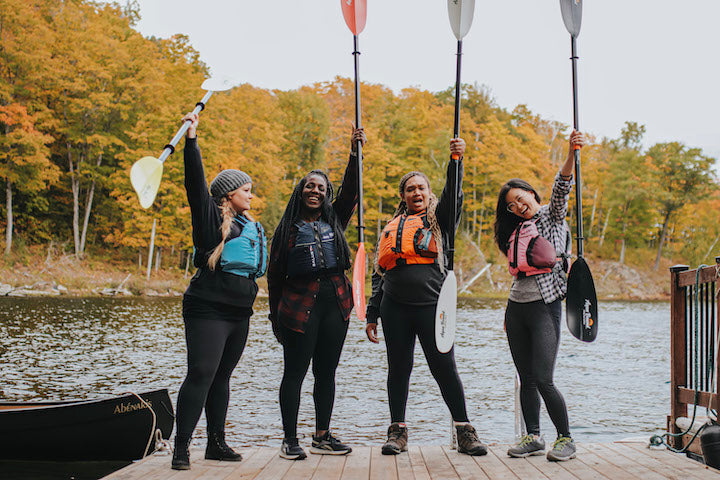 four young women stand on a dock raising kayak paddles up in the air