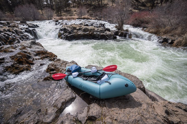 packraft sits next to a whitewater river equipped with paddle/pogies and other ultralite gear