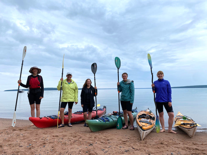 5 women stand next to four kayaks, each holding a paddle, on a sandy beach