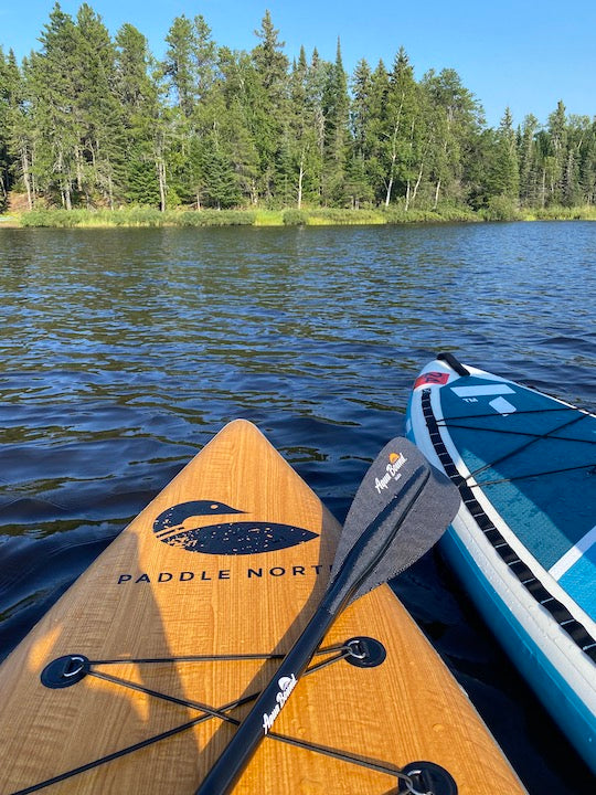 noses of two paddleboards, with a paddle on a lake