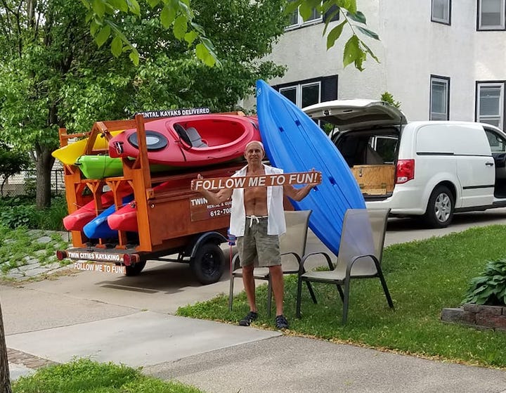 Twin Cities Kayaking owner, John Schulte, with his van and trailer full of kayaks; holding a sign that says "follow me to fun"