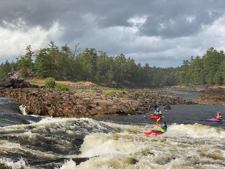 three kayakers in river rapids, views of rock bar and trees on shoreline