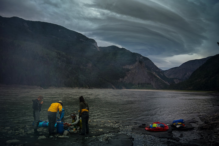 three people on shore with gear next to the river, stormy clouds overhead