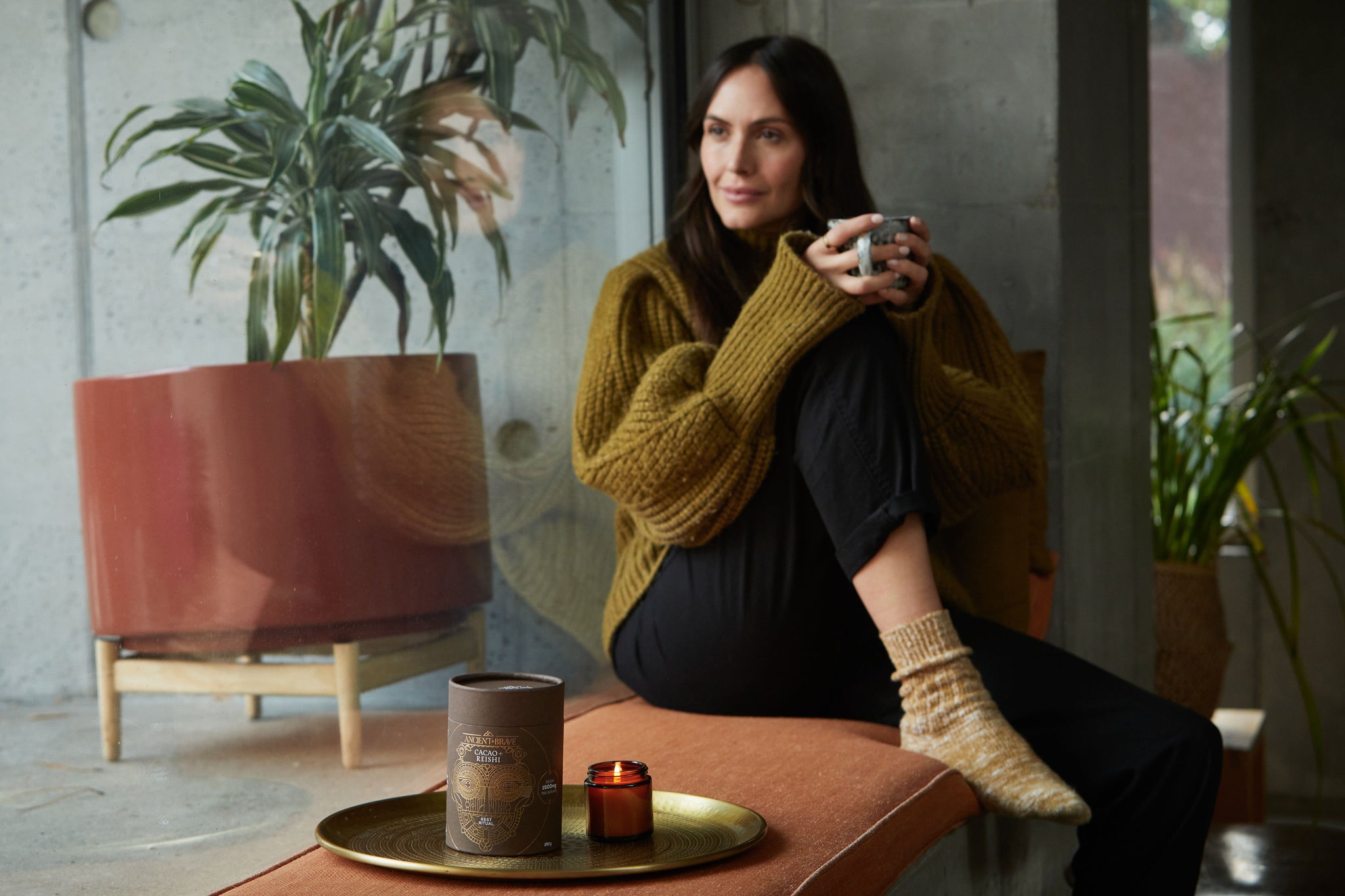 Image of a woman in a yellow knit jumper sat on a window seat looking outside. Holding a mug of Cacao + Collagen.