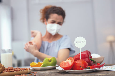 Frau kratzt sich am Arm, davor ein Teller mit Gemüse und ein Schild " Allergie"