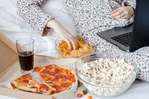 Gesunde Snacks für abends: Person liegt mit Laptop im Bett, daneben Pizza, Chips, Popcorn und Cola