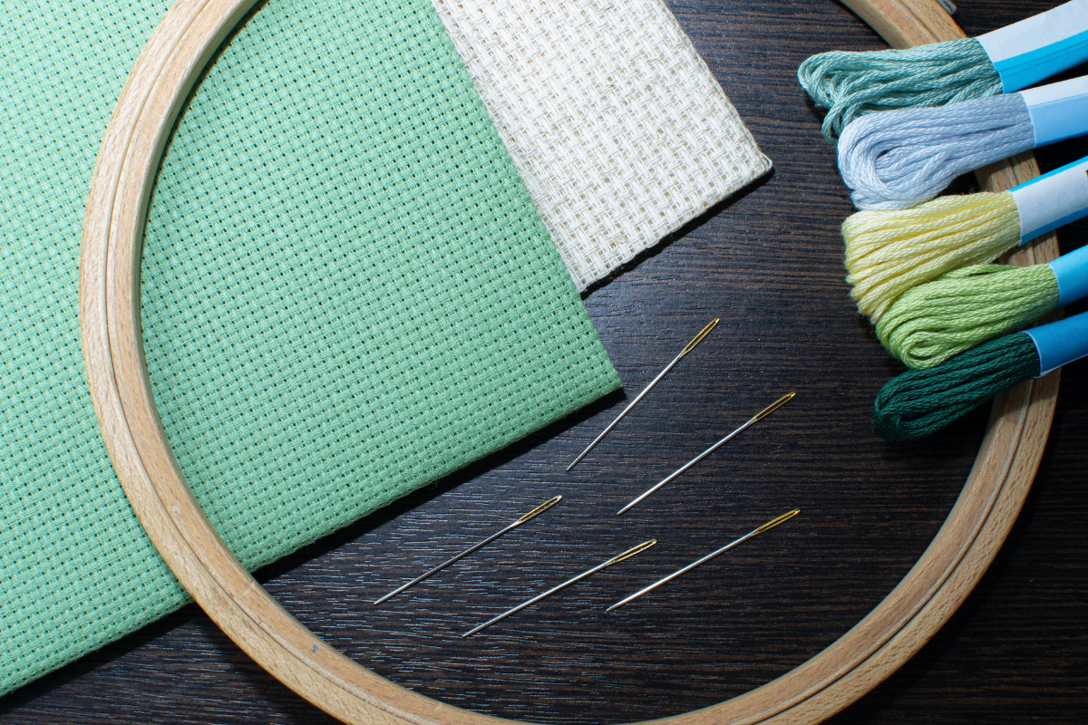 cross stitch fabric in white and green with wood embroidery hoop, several sets of embroidery thread and cross stitch needles