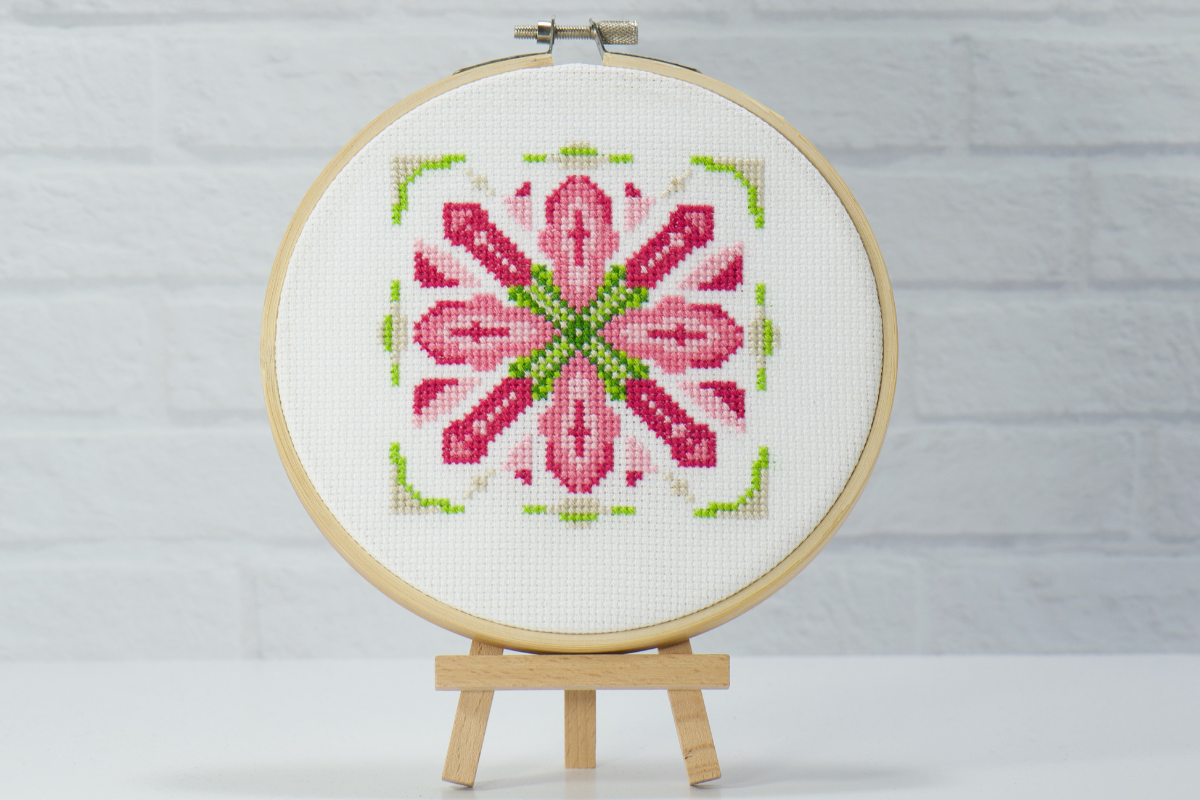 pink and rose cross stitches into a flower with green center surrounded by softer green and beige embroidery stitches on white cross stitch fabric inside embroidery hoop