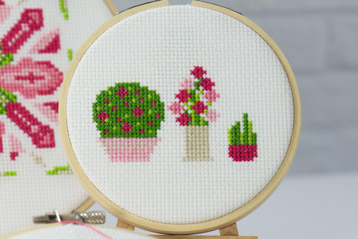 three potted flowers cross stitched with vibrant floss on white cross stitch fabric inside wood embroidery hoop.