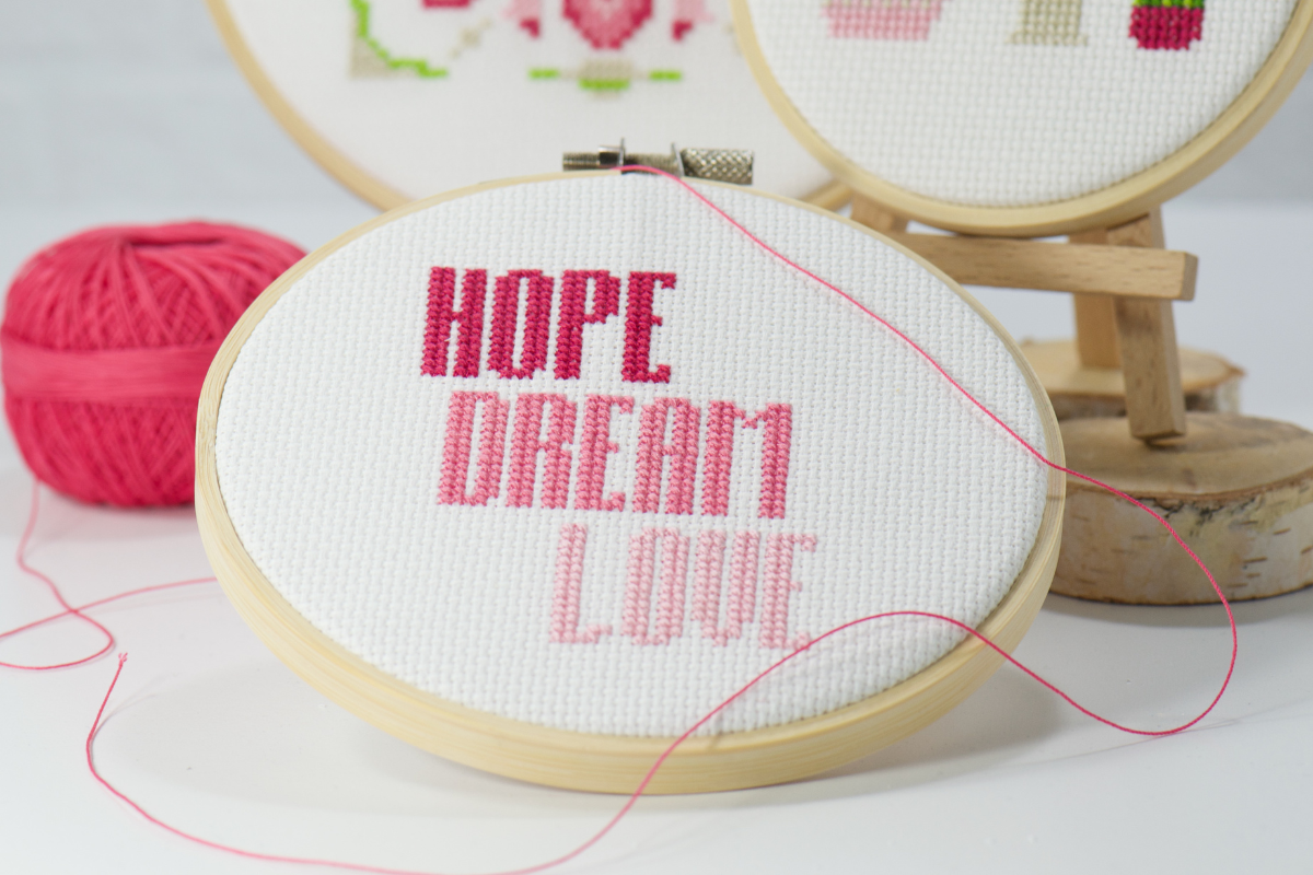 cross stitch words in three shades of pink on white cross stitch fabric inside wood embroidery hoop