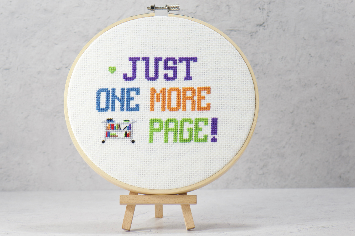 book lovers cross stitch pattern featuring just one more page words with book cart on white cross stitch fabric inside wood embroidery hoop