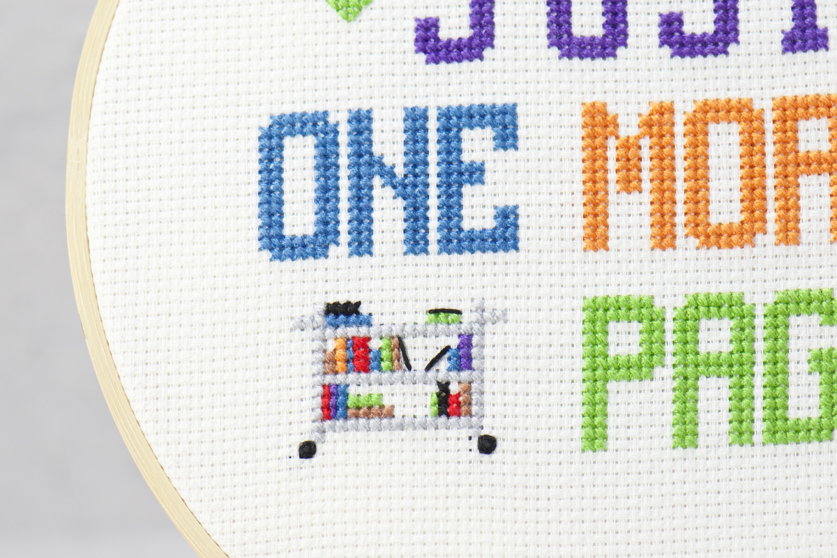 just one more page book lovers cross stitch design with words and a book library cart on white cross stitch fabric