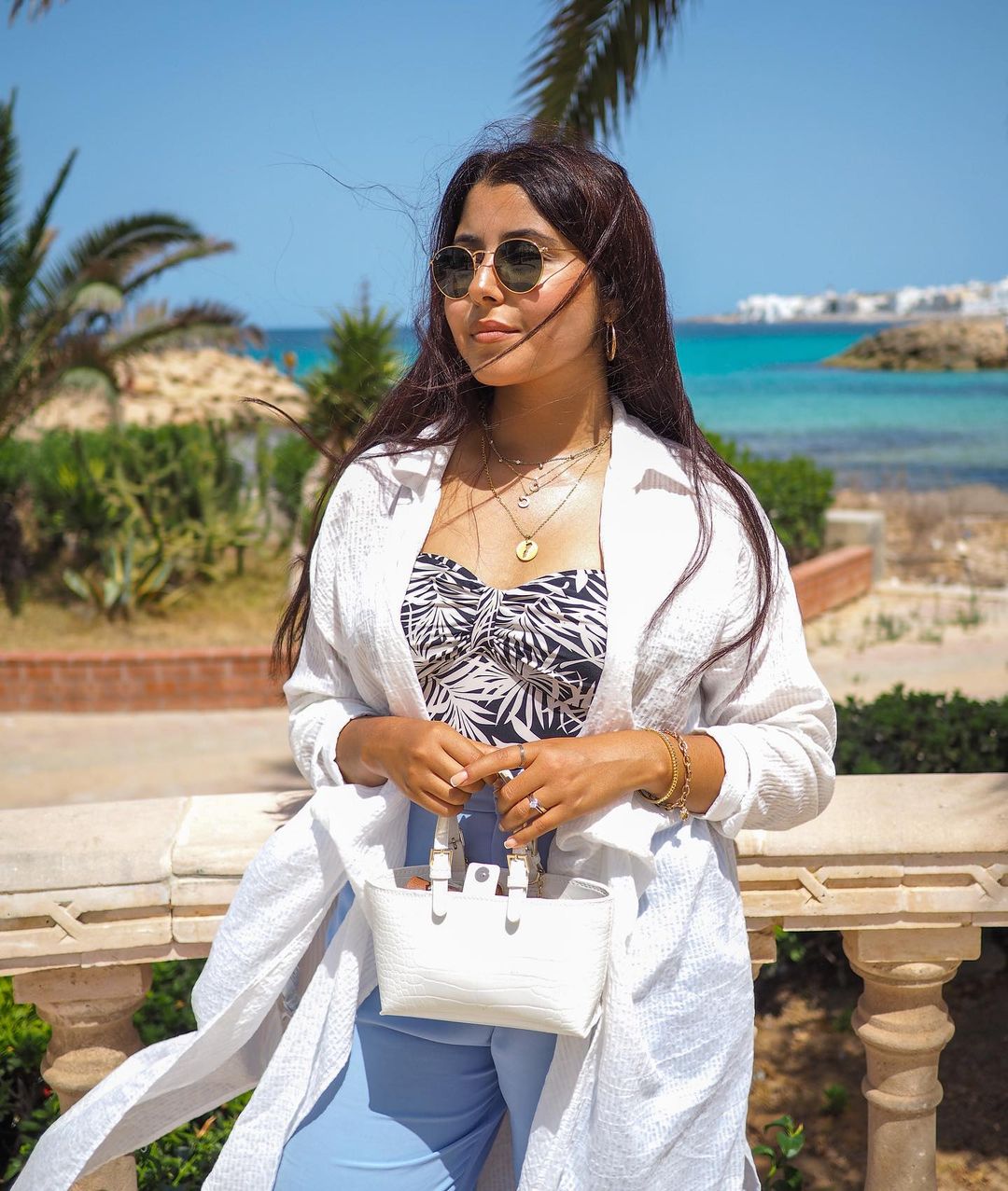 Girl on a beach blue sky ocean sand with a stack of gold necklaces layered with sunglasses and long hair.