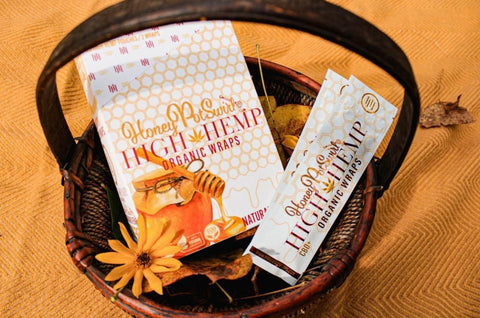 Square image with a brown wooden basket sitting against an orange background.  The basket holds one single Honey Pot Swirl High Hemp Wrap package, a full box of the same Honey Pot Swirl High Hemp Wraps, and a single orange daisy.