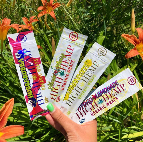 Four different flavors of High Hemp Wraps - Hubba Bubba, Hydro Lemonade, BananaGoo, and Blazin' Cherry - being held by a hand in front of a background of orange lilies and their long green stems.  The visible thumbnail on the hand is painted seafoam green.