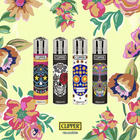 square image with light yellow background with blue, pink, purple and green flowers along the border.  Centered in the photo are four Clipper lighters, each with a different sugar skull design.  Each lighter is also a different color.  From left to right, the lighters are as follows:  Horizontal stripes in pink and mango with a black skull that features light blue and magenta flowers, a white nose, yellow stars for eyes, teeth are barely visible thin, white hatching similar to a luchador's mask; solid black background with white skull featuring grey flowers and hearts, one eye is a red rose and one a grey flower, the nose is black, the teeth are white and smiling wide; a white background with yellow and black flowers featuring a blue skull with orange and yellow flowers, big, straight, white teeth, a blue nose, and two orange and yellow flower eyes; and a solid black background featuring a multi colored skull of pink, blue, green, and yellow geometric shapes, black eyes, and a medium white  half smile.
