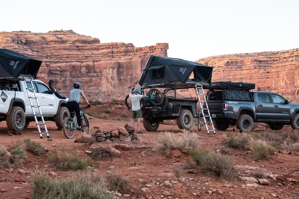 Two campers riding bikes in the desert in front of Toyota Tacoma with Skycamp 3.0 and Toyota Tacoma with Skycamp 3.0 mounted over trailer rack. 